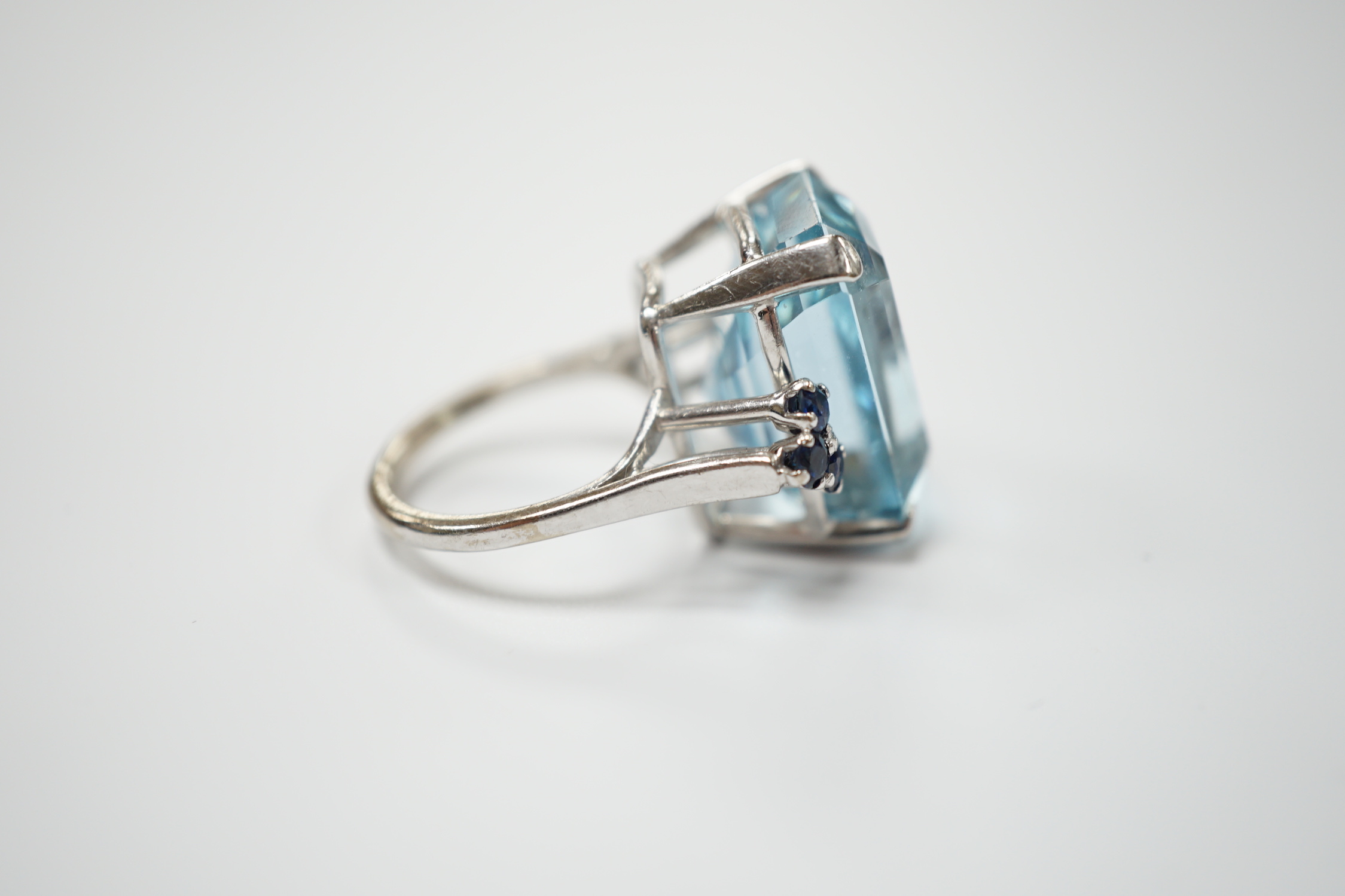 An 18ct white metal and single stone emerald cut aquamarine set dress ring, with six stone sapphire set shoulders, size L, gross weight 11.8 grams.